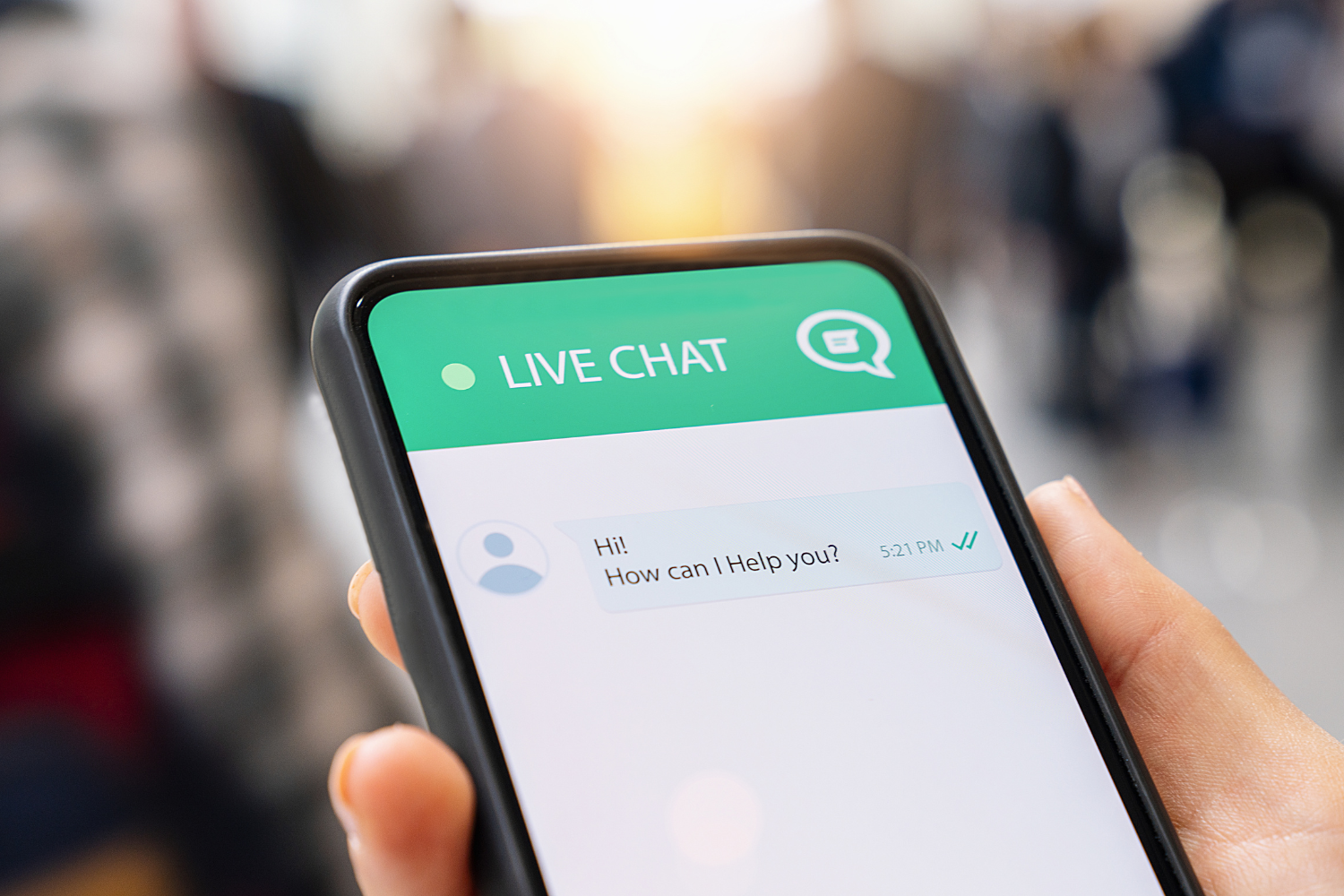 A persons hand holding a smartphone with "Live Chat" in a green banner across the screen. The chat says "Hello! How can I help you?"